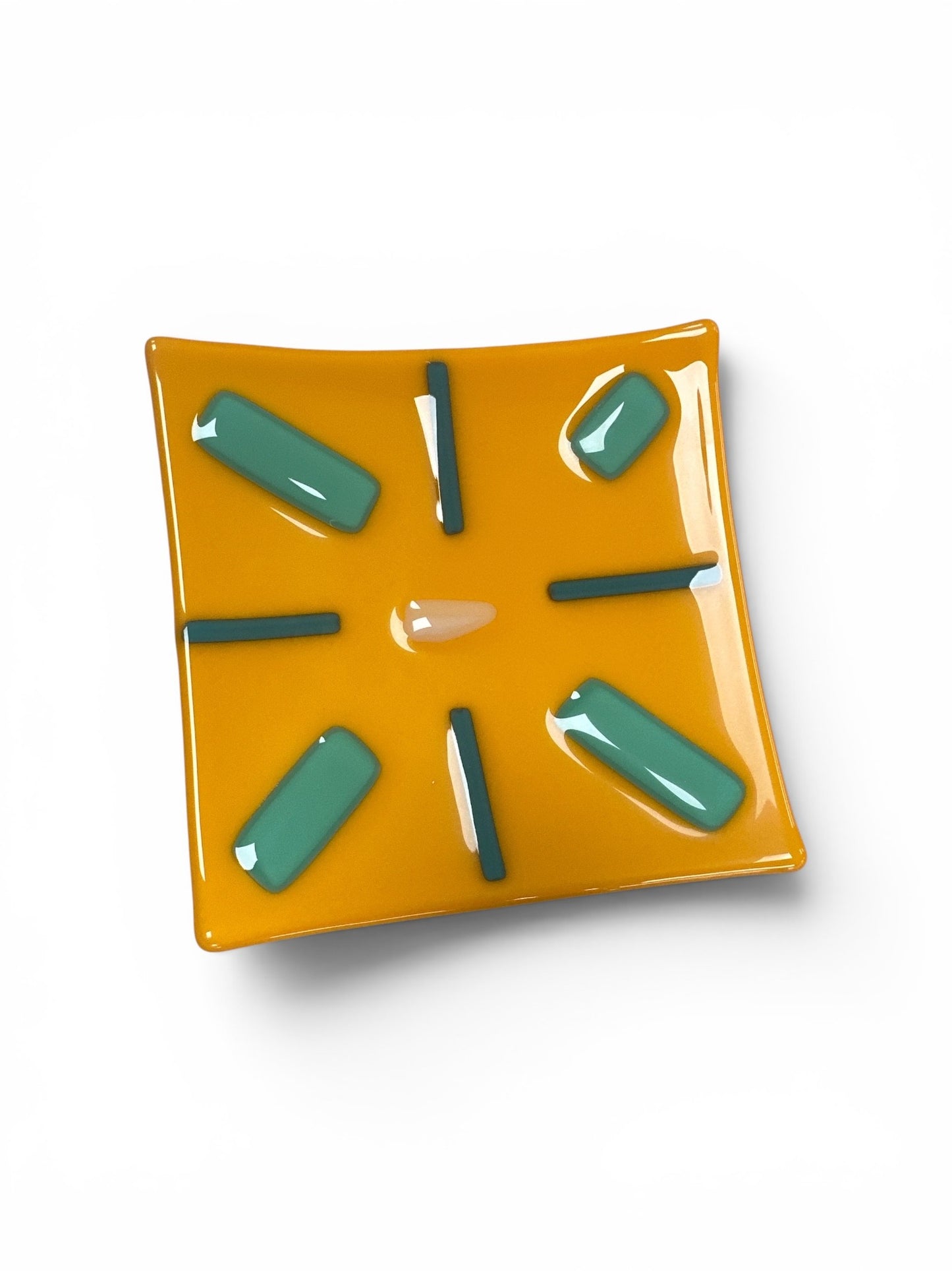 This is a square, mustard yellow fused-glass decorative dish featuring a central beige accent surrounded by a geometric, starburst-like pattern of teal bars and thin, dark gray glass lines.
Medium: Fused Glass
Size: Approximately 6" x 6"
Year: 2026