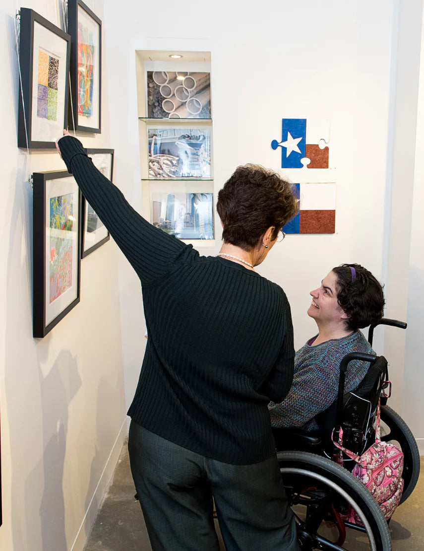 A female with brown hair in a wheelchair next to a woman standing and pointing at artwork on the wall.