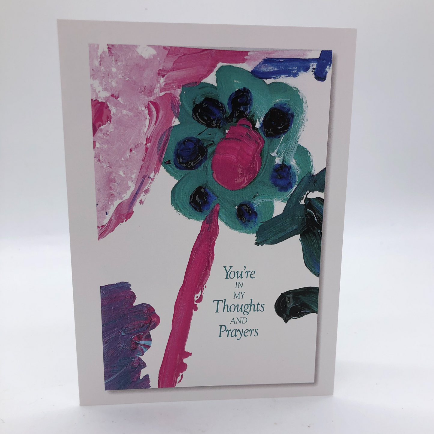 The greeting card has a white background and is printed with a picture painted with acrylics. The focus of the card is a teal and dark blue flower with a magenta center on a magenta stem. The upper left of the card has a wash of magenta in a triangular shape. The lower left has a purply swash of color and there are some strokes of the dark blue on the right hand side middle. In the lower right are the words "you're in my thoughts and prayers."