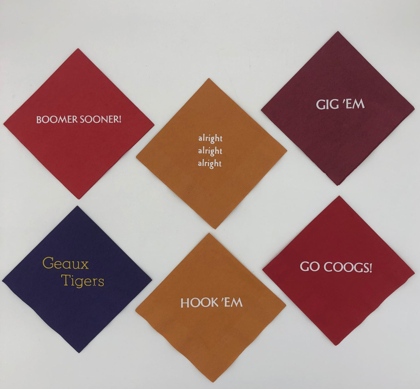 Varying colored napkins with college slogans including red napkin with Go Coogs! slogan, burnt orange napkins with Hook 'Em and alright, alright, alright slogans., purple napkin with gold Geaux Tigers slogan, red napkin with white Boomer Sooner! slogan, and maroon napkin with white Gig 'Em slogan