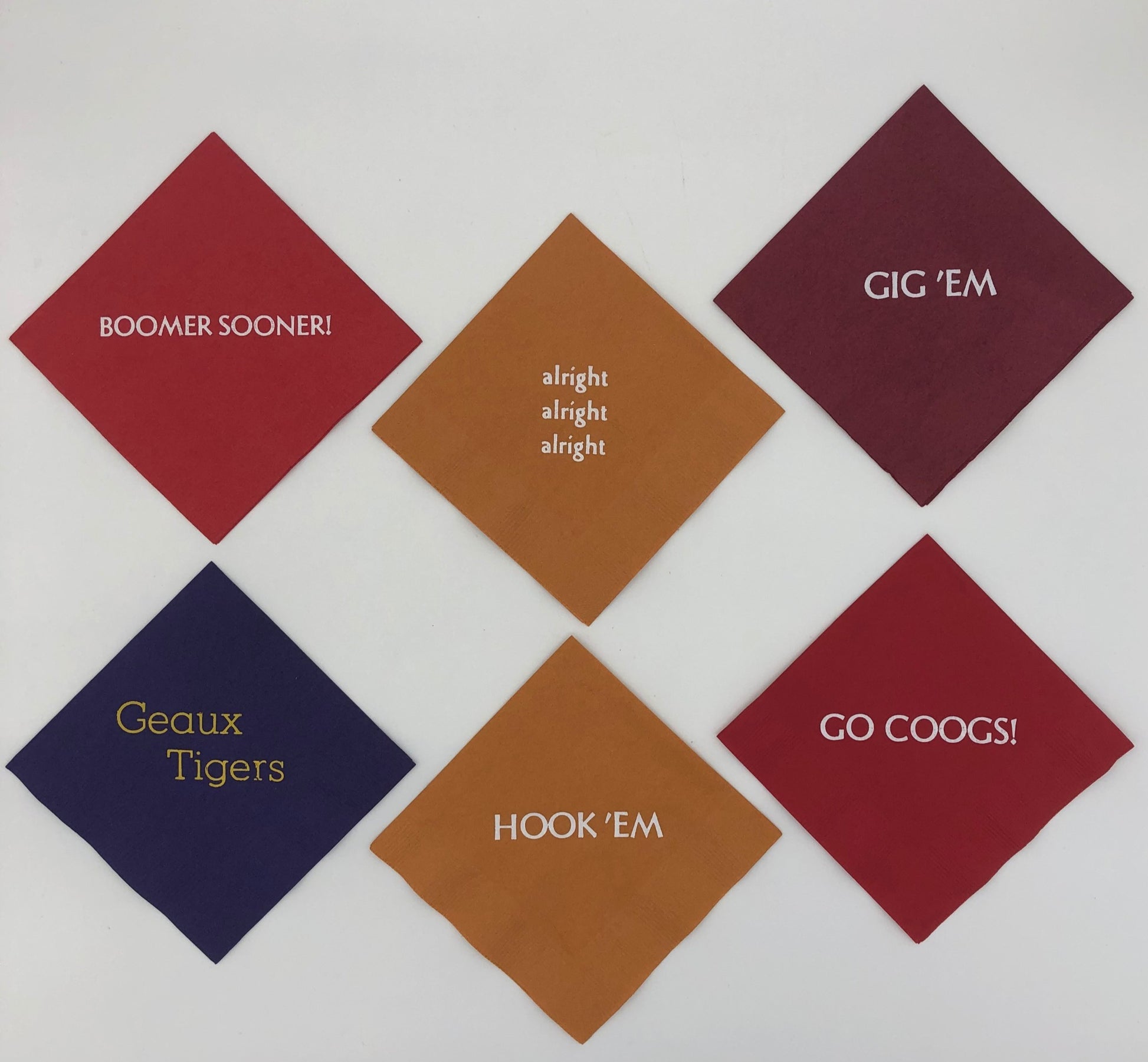 Varying colored napkins with college slogans including red napkin with Go Coogs! slogan, burnt orange napkins with Hook 'Em and alright, alright, alright slogans., purple napkin with gold Geaux Tigers slogan, red napkin with white Boomer Sooner! slogan, and maroon napkin with white Gig 'Em slogan