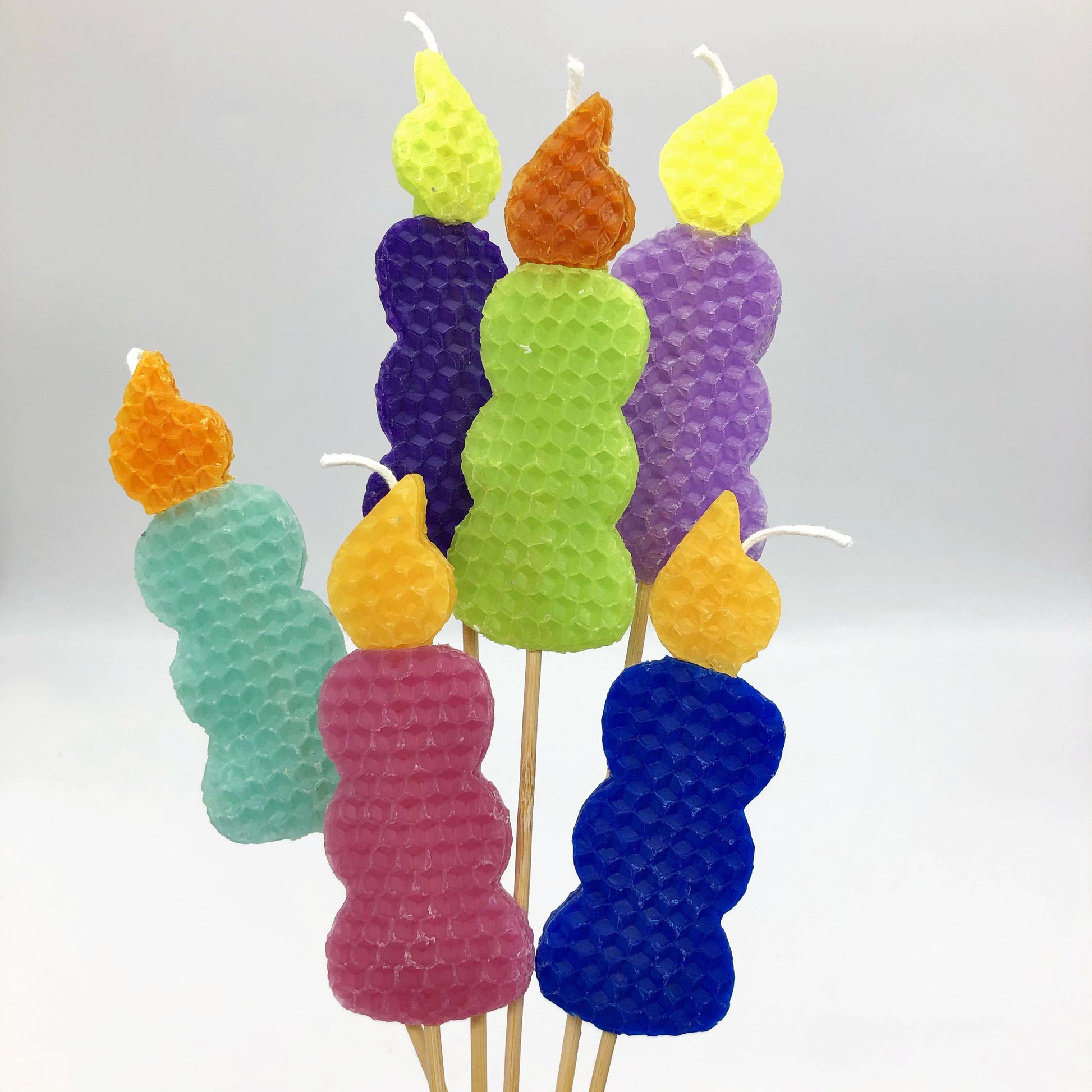 Beeswax candles in the shape of a candle with yellow and orange flames with blue, pink, green or purple bases