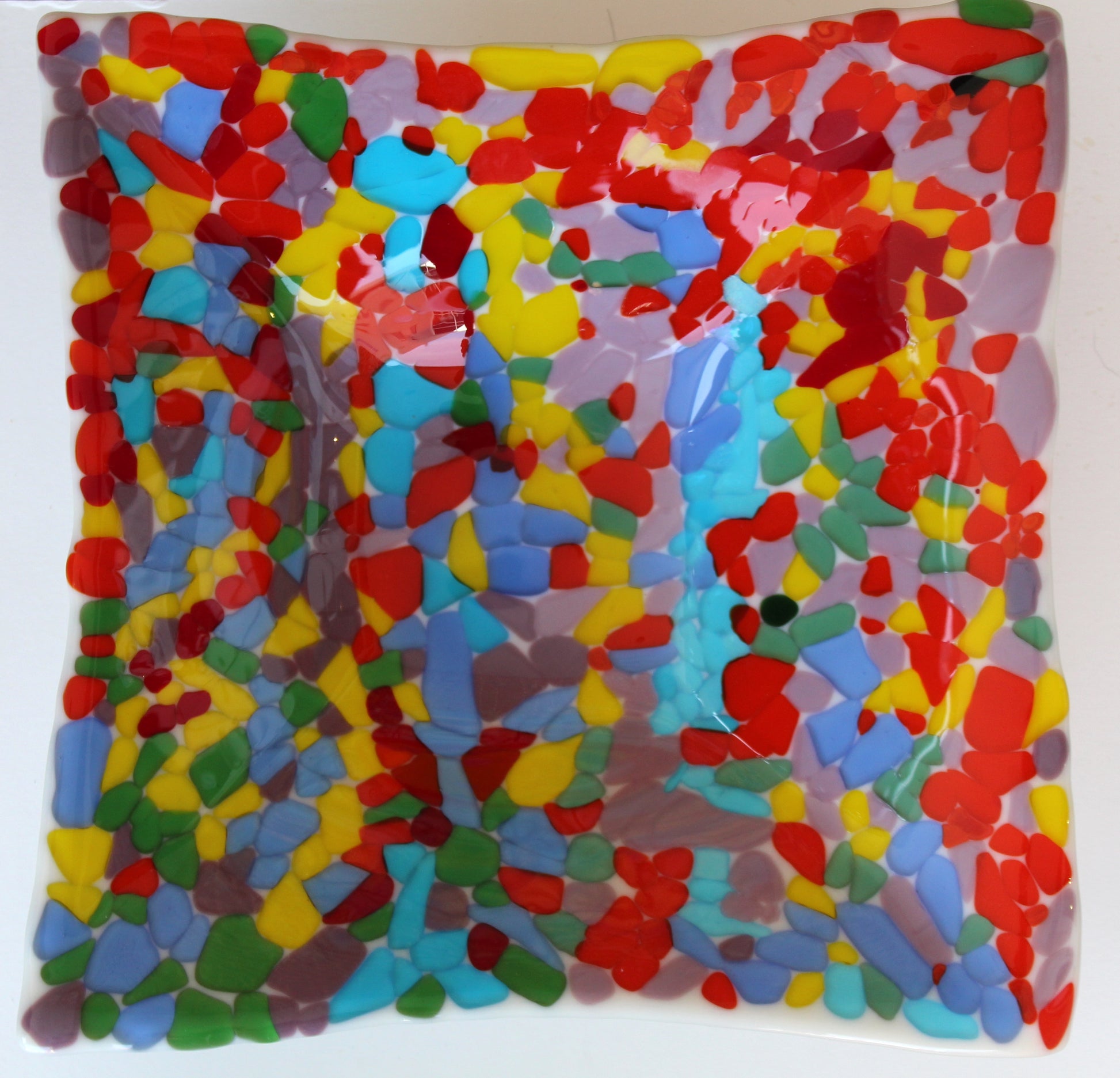 White glass square bowl completely covered in transparent chunks of bright colorful glass of reds, yellow, blues, green, and purple