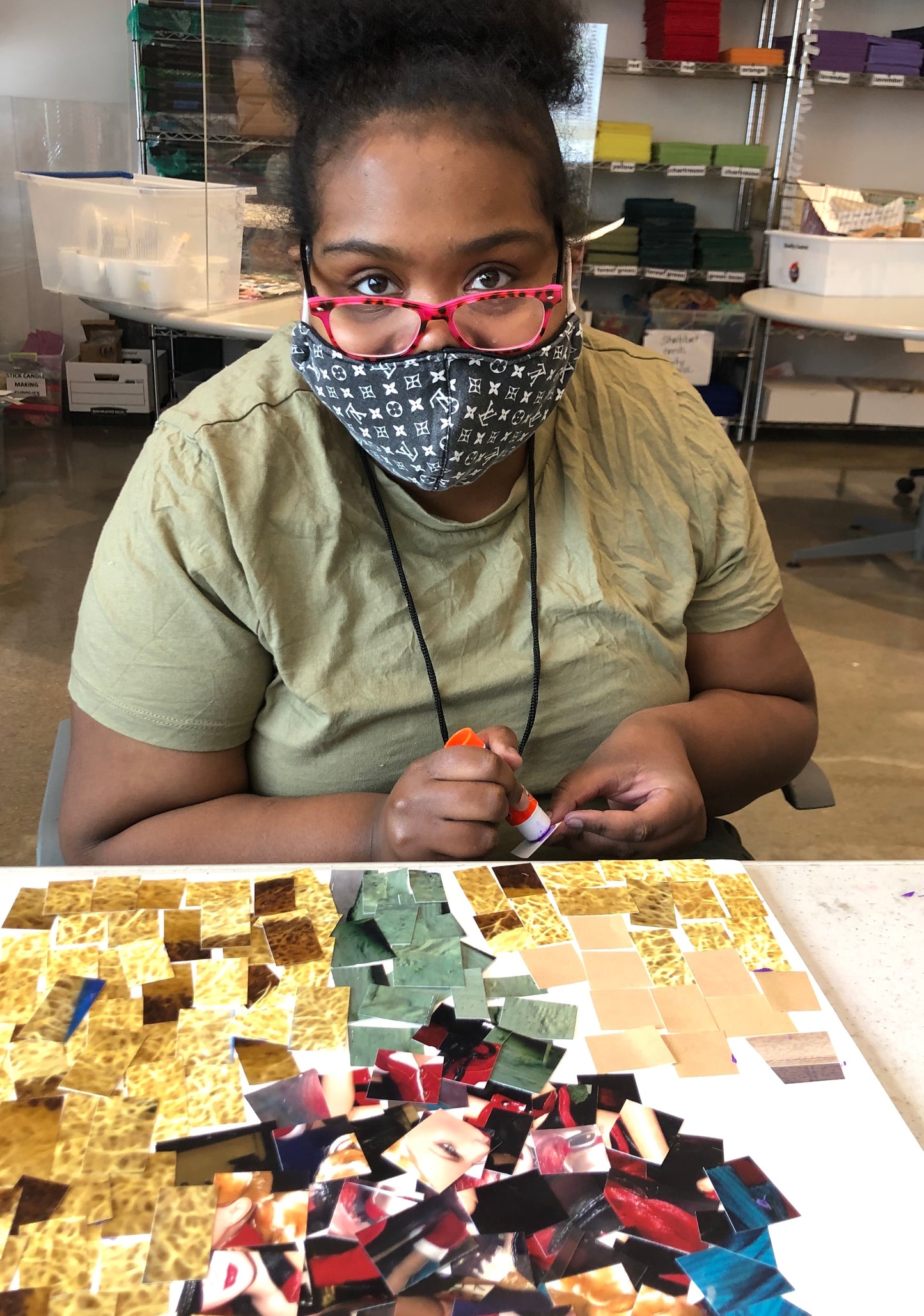 Female participant with pink eye glasses looking toward the camera and glueing a piece of her mosaic art.  Mosaic art is sitting on the table in front of her.  It is made of of square of photographs in shades of cream, yellow, red and green.