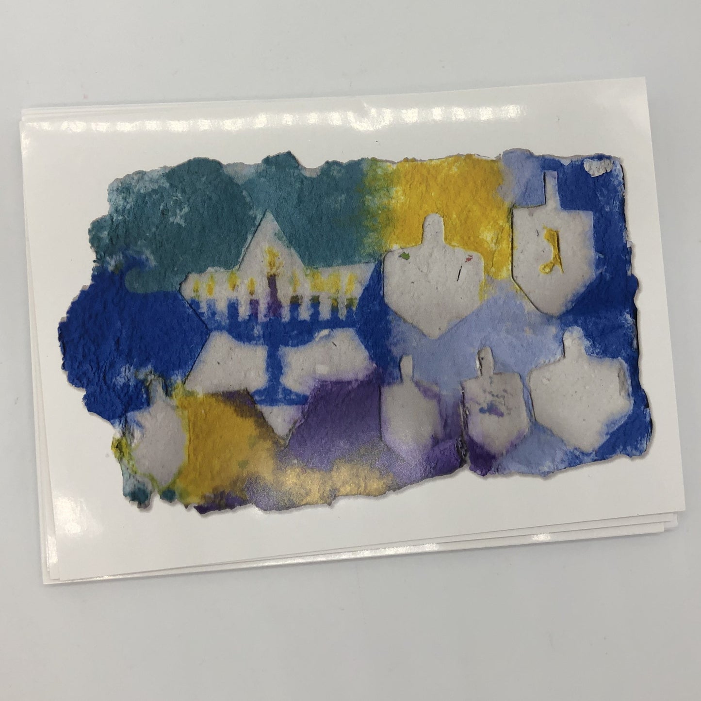 Photograph of handmade paper in shades of blue, teal and yellow. There is one large Star of David with a blue menorah inside and several white dreidels.