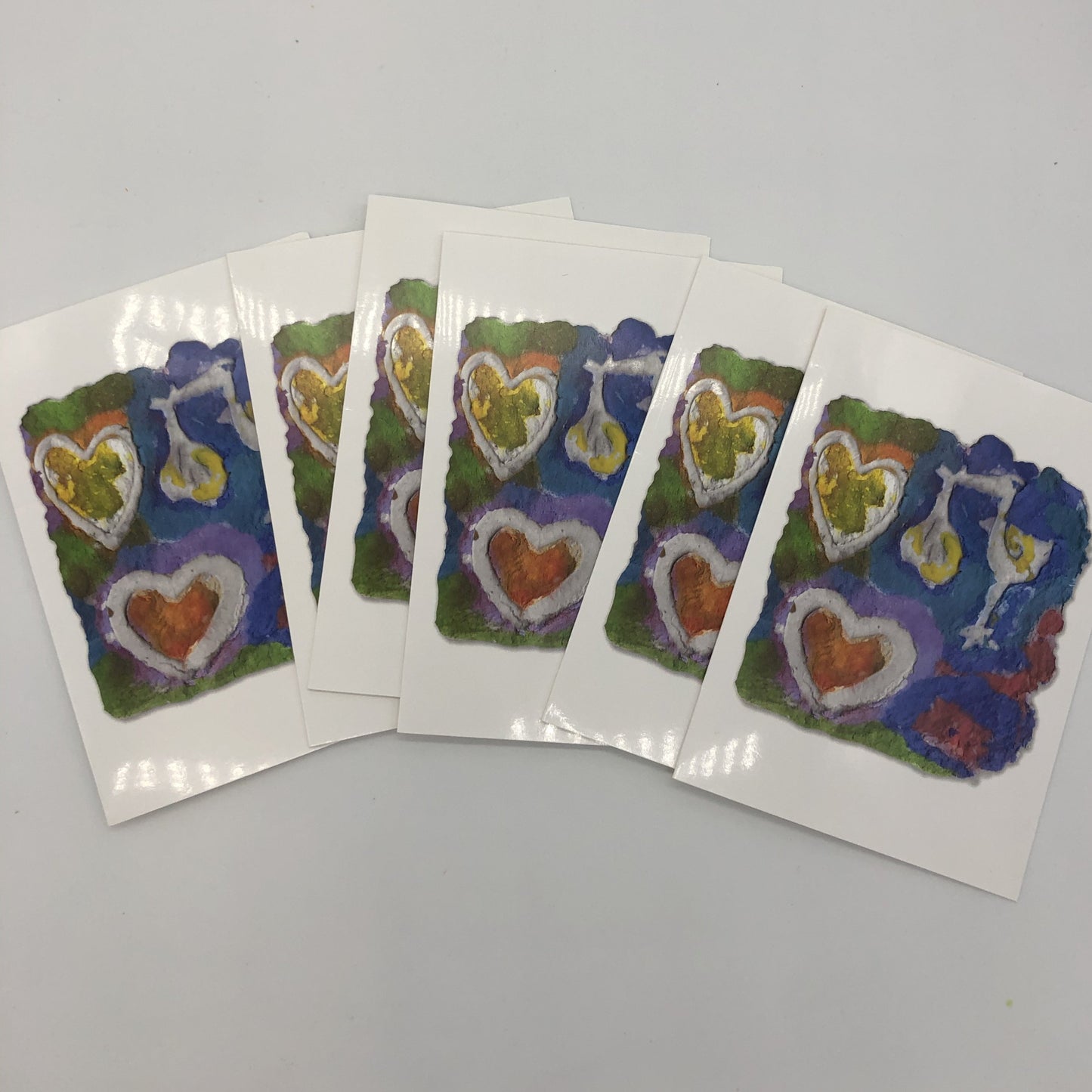 Six cards fanned out. Printed card of photo of handmade paper in blues, greens and reds. On top of the design are two white hearts and a stork holding a wrapped baby by it's beak.