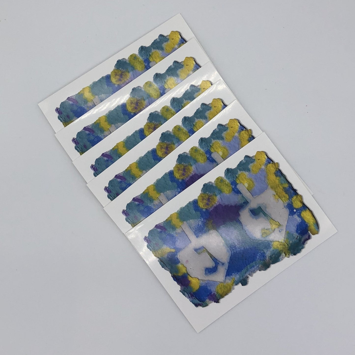 Six of the greeting cards fanned out. White greeting card with the printed image of handmade paper in shades of yellow, blue and a little bit of purple.  There are two large dreidels side by side with the Hebrew symbol