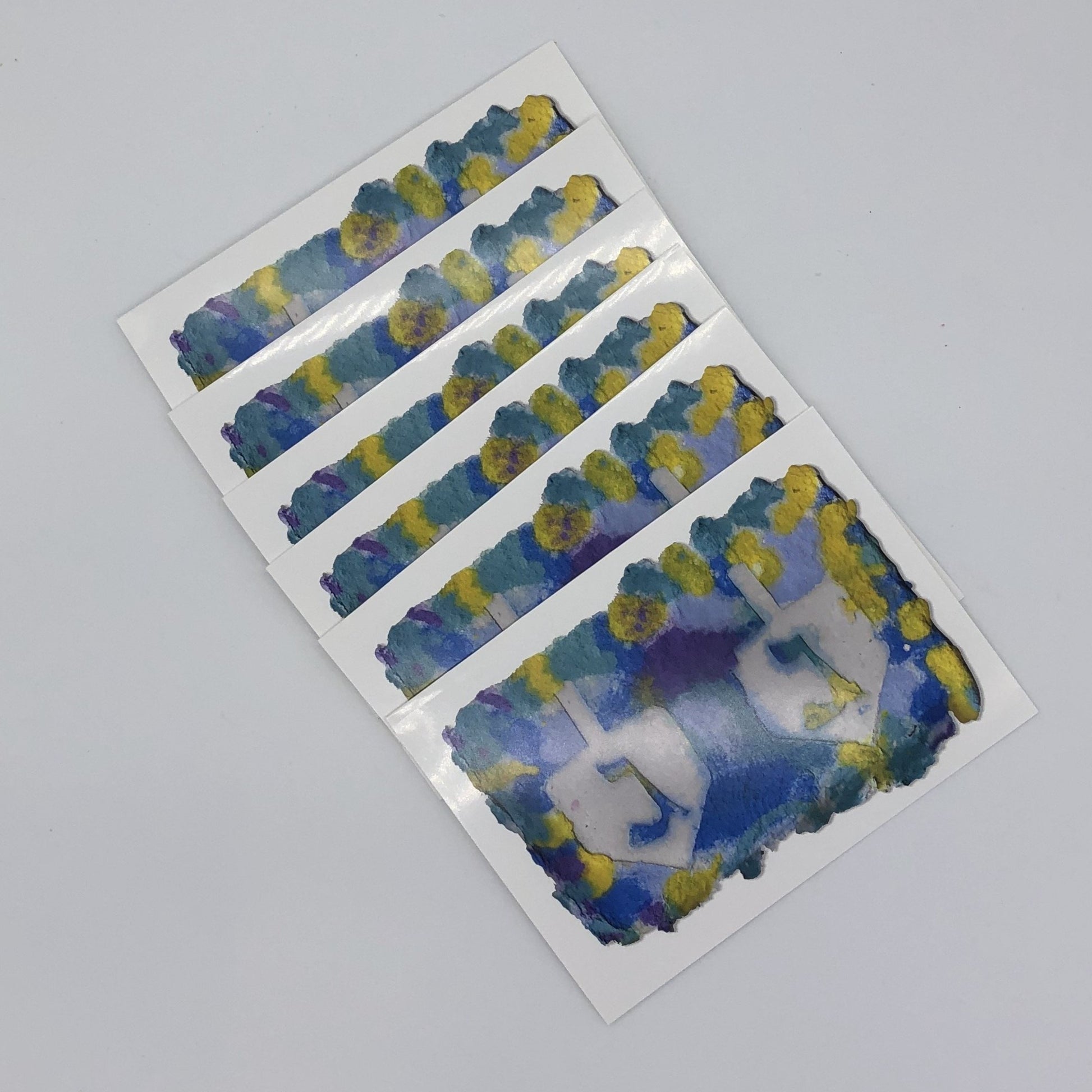 Six of the greeting cards fanned out. White greeting card with the printed image of handmade paper in shades of yellow, blue and a little bit of purple.  There are two large dreidels side by side with the Hebrew symbol