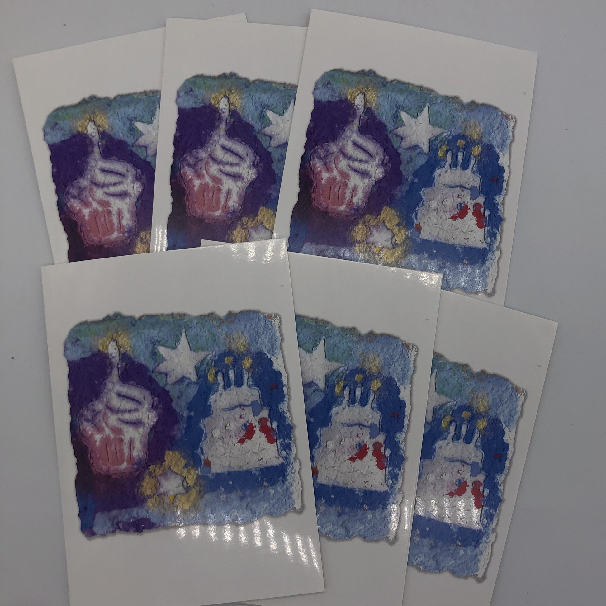 Six of the birthday cards fanned out. Printed card of a picture of handmade paper in blues and purple with a little gold. On top of the paper are stars, cupcake and cake with candles.