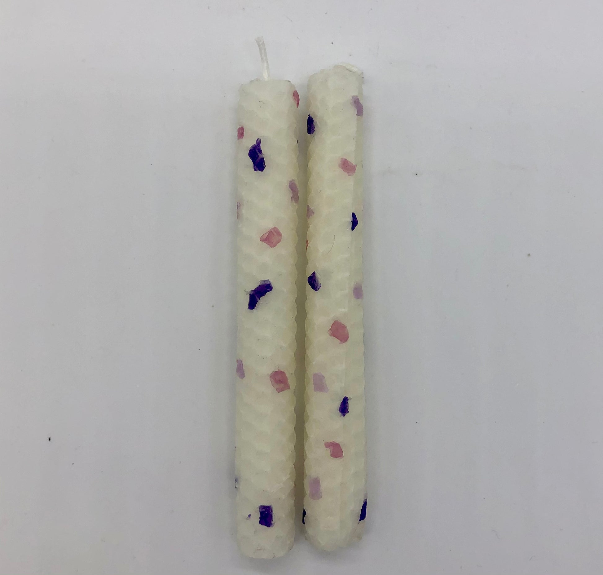 Two white shabbat candles with dots of color in purples and pink.
