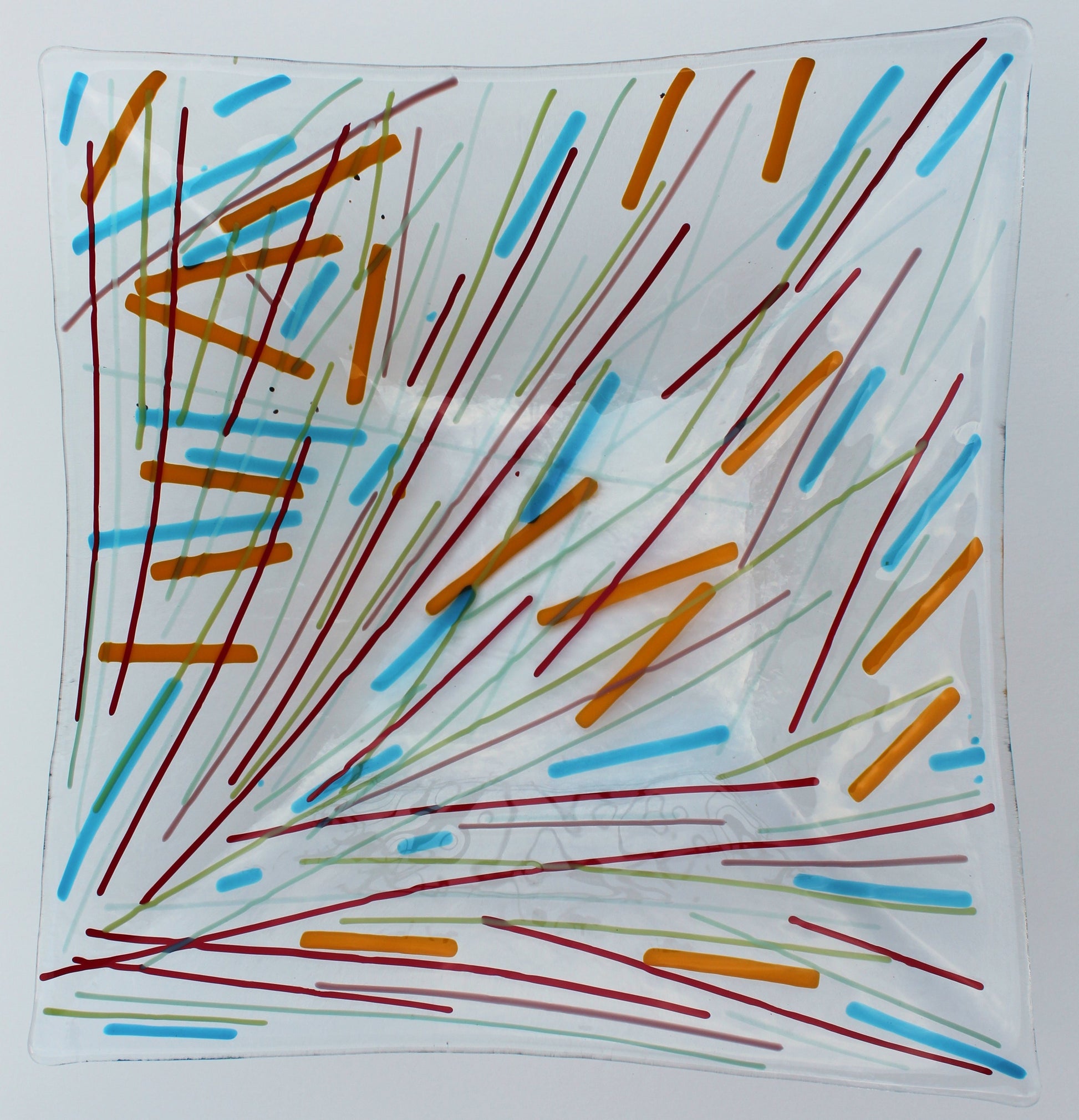 Fused glass square bowl. Bowl is a made of a clear piece of glass with lines radiating from the lower left corner outward. The colors of the lines are red, green, faint blue, with thicker lines of orange and light blue.