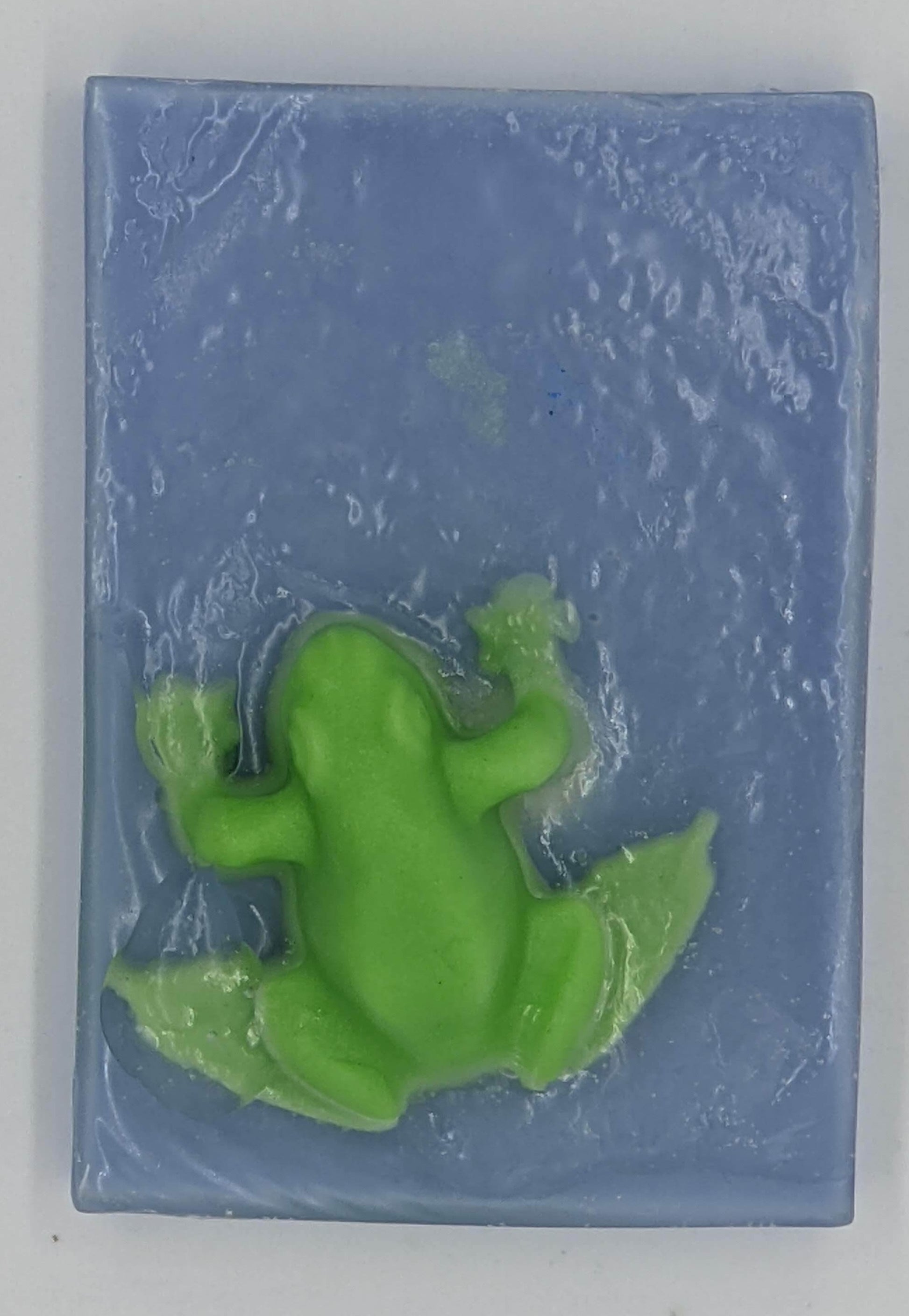 Bar soap with a green frog protruding. There is a thin layer of blue, then clear, and the green frog is embedded in the clear portion to appear as is the frog is sitting on top of water