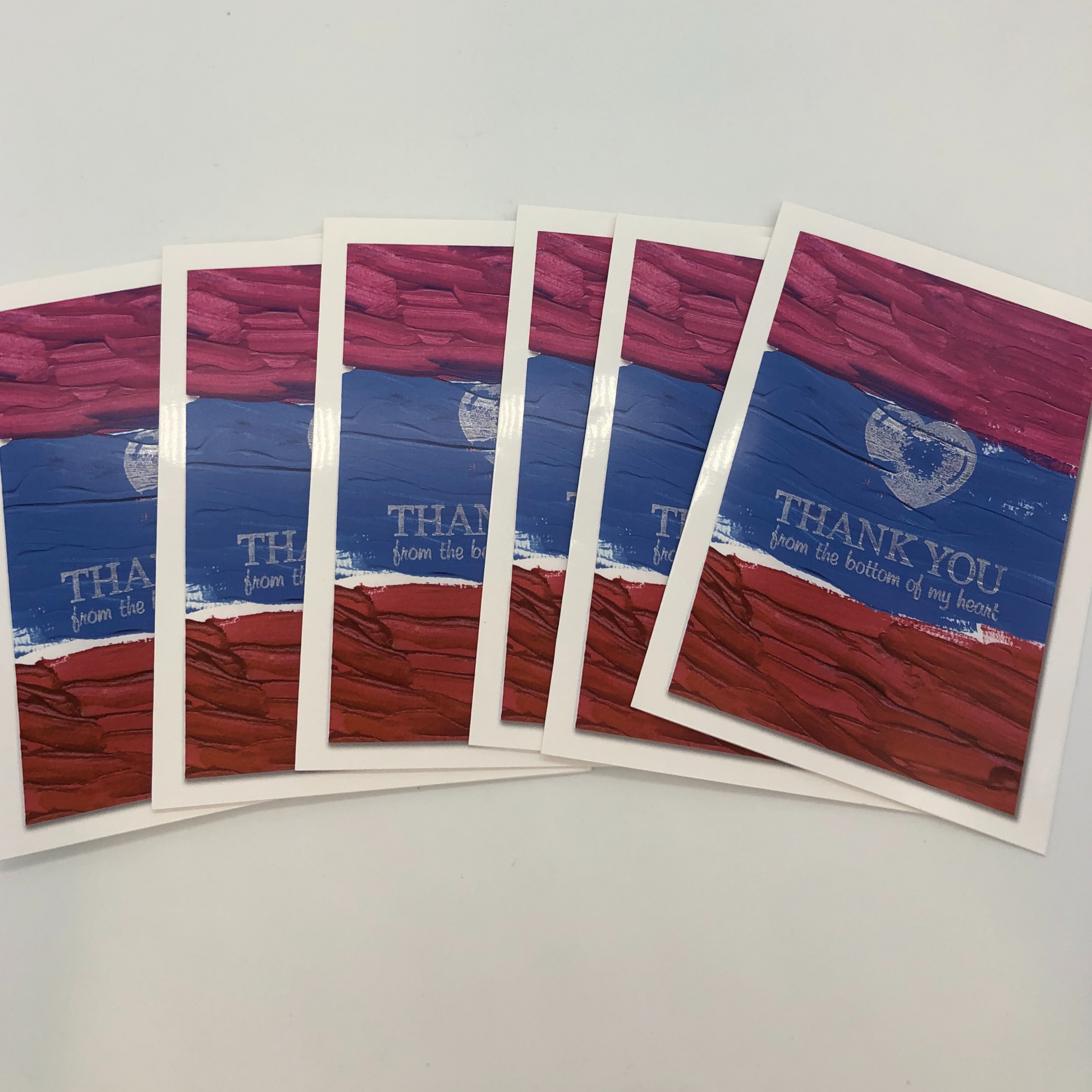 Photo showing that six cards and envelopes come in each packet.  Each card is a print of an acrylic painting with stripes of three colors-maroon on top, bluish purple in the middle and red on bottom.  Over the top is a white stamp of a heart and the words Thank you from the bottom of our heart.