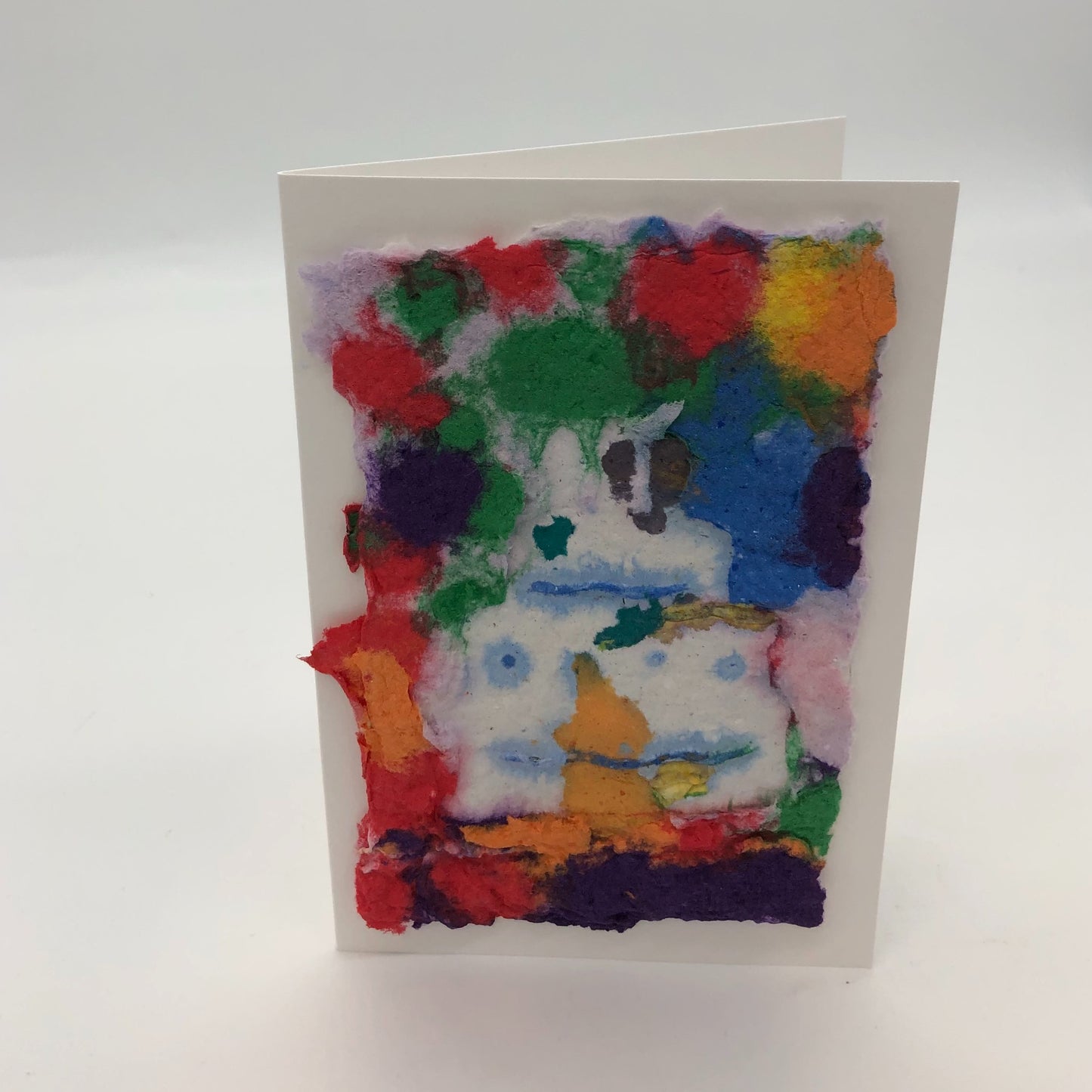 Handmade paper with white cakewith light blue details on top. Background is blobs of several different colors including red, green, blue, purple, orange and yellow.