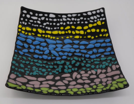 A black square glass curved plate that has small pebbles of colors. There are on average of three rows each color before transitioning to the next color. Colored pebbles are placed in the following order: white, yellow, blue, turquoise, light purple, and green.