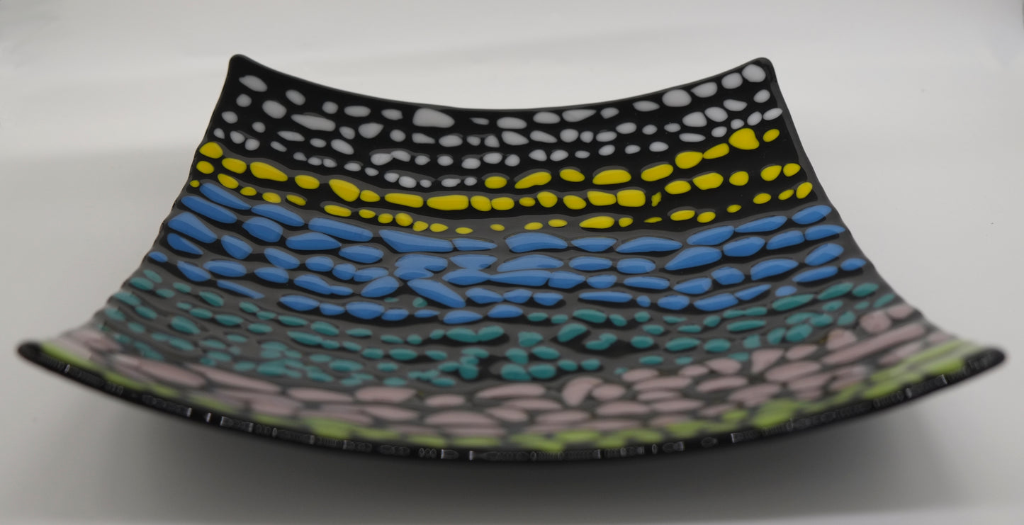 A black square glass curved plate that has small pebbles of colors. There are on average of three rows each color before transitioning to the next color. Colored pebbles are placed in the following order: white, yellow, blue, turquoise, light purple, and green.