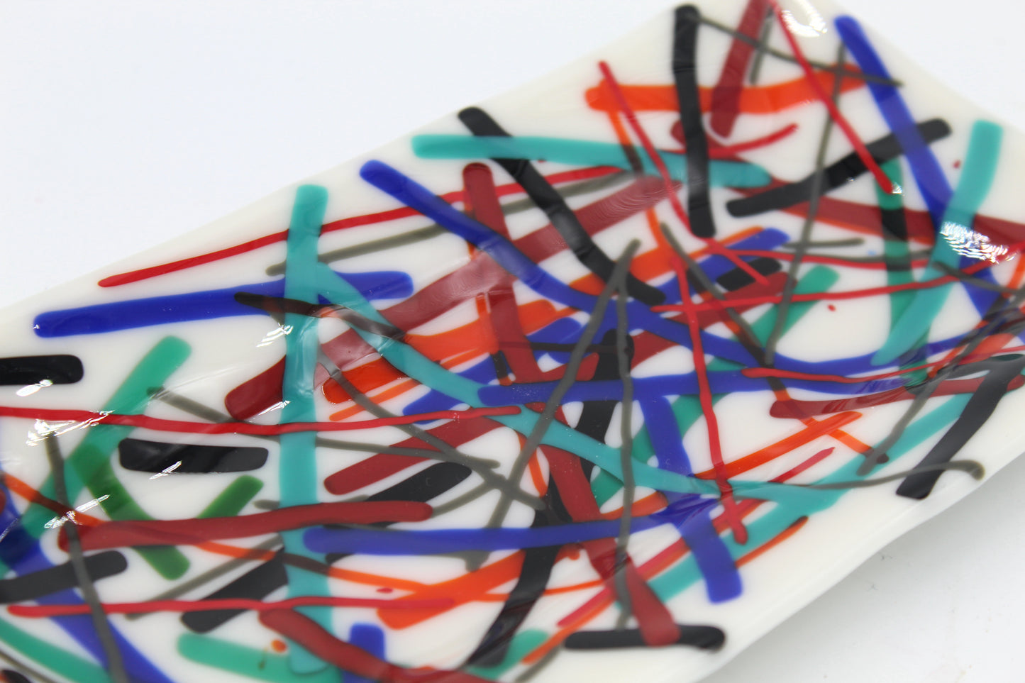 This is a piece of fused glass that is white with muticolored linked in abstarct form. Colors include: black, blue, light blue, red, and orange
