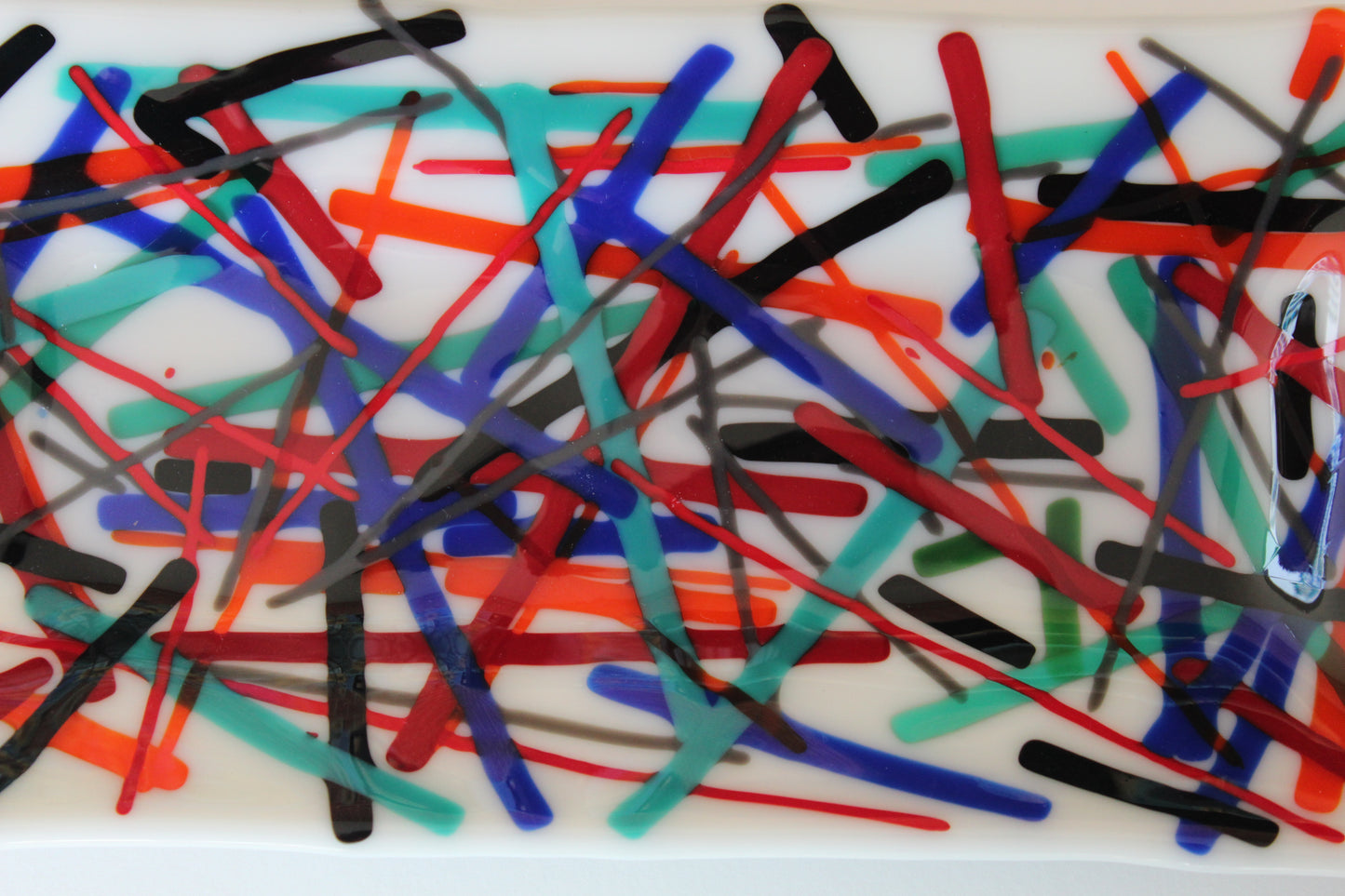 This is a piece of fused glass that is white with muticolored linked in abstarct form. Colors include: black, blue, light blue, red, and orange