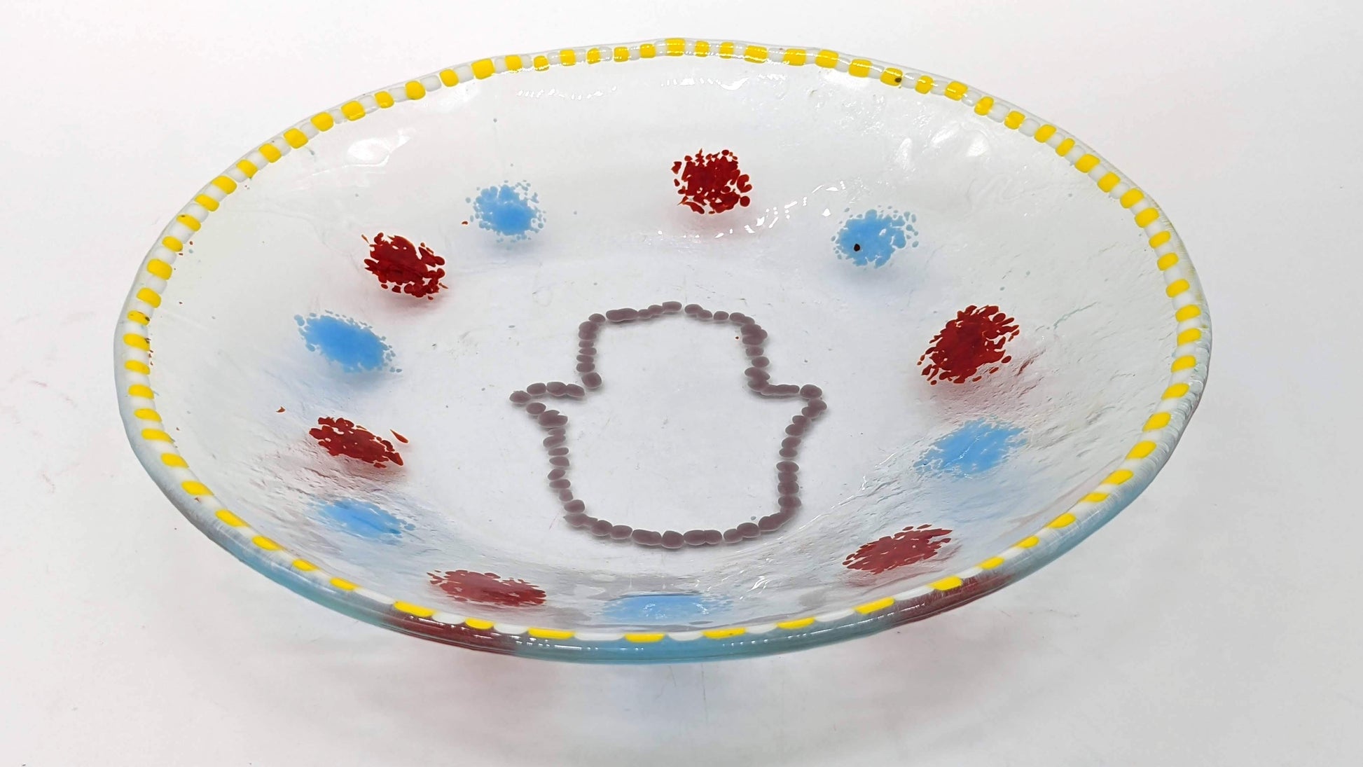 clear glass bowl with an outline of a hamsa in the center surrounded by alternating dots of blue and red with a border of yellow and white