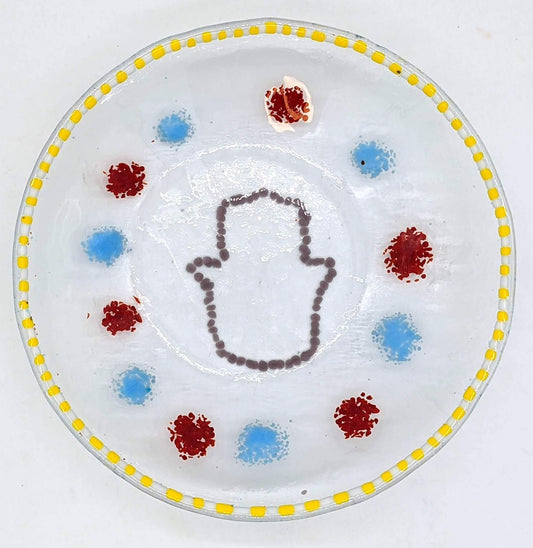 clear glass bowl with an outline of a hamsa in the center surrounded by alternating dots of blue and red with a border of yellow and white