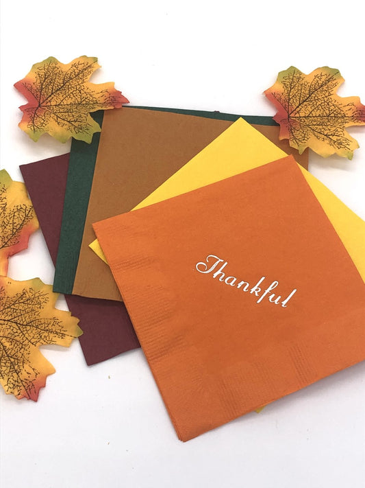 Cocktail napkins in maroon, dark green, light brown, yellow and orange.  The orange napkin has a silver Thankful slogan