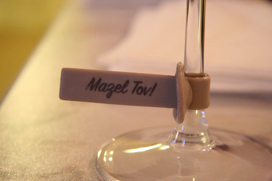 A close-up of a wine glass stem with a beige tag attached that reads "Mazel Tov!" in black script. 