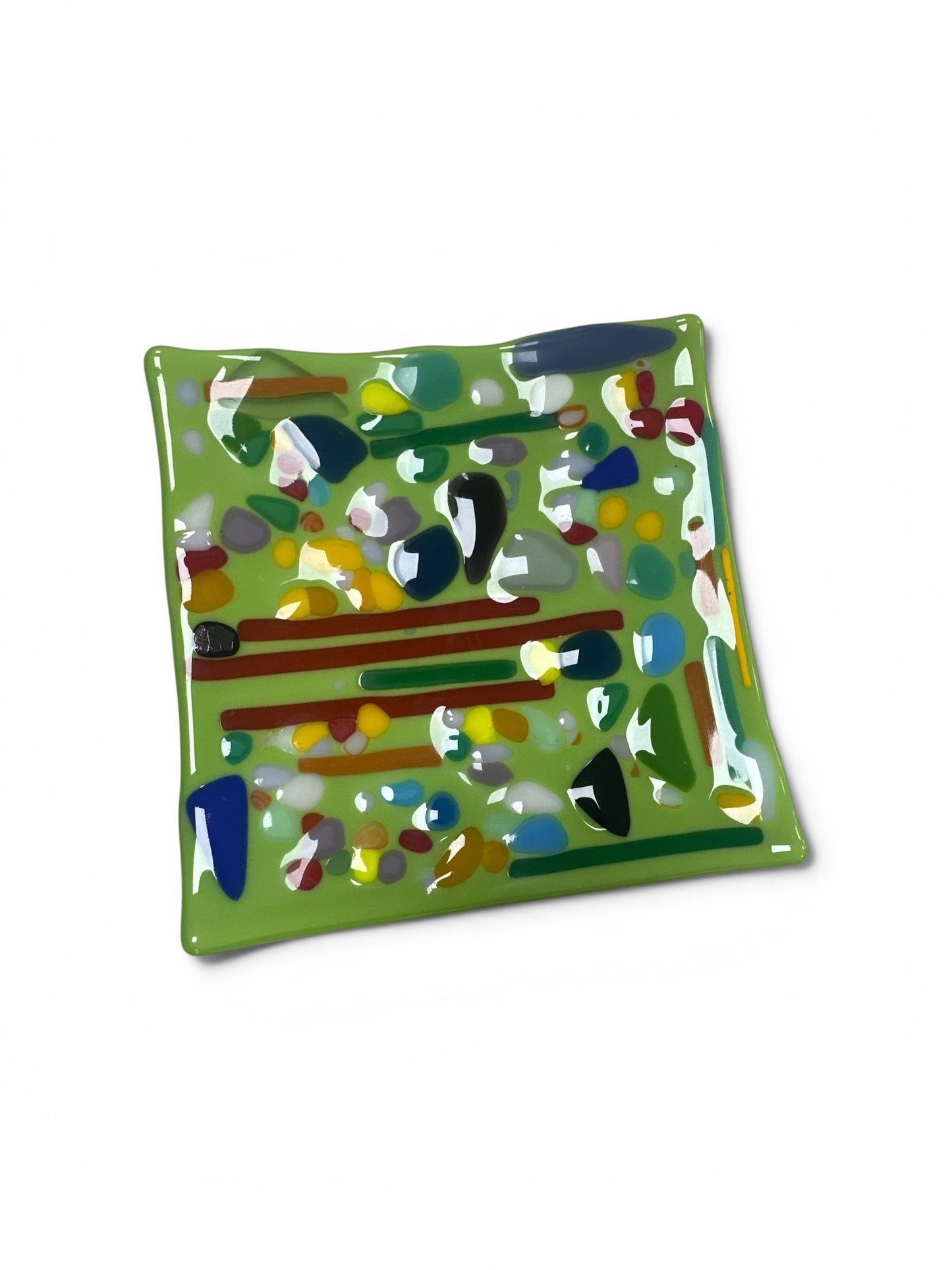This is a square, light green fused-glass decorative dish covered in a vibrant, collage-like mosaic of multi-colored glass pebbles, shards, and thin stripes in shades of red, yellow, blue, and dark green.