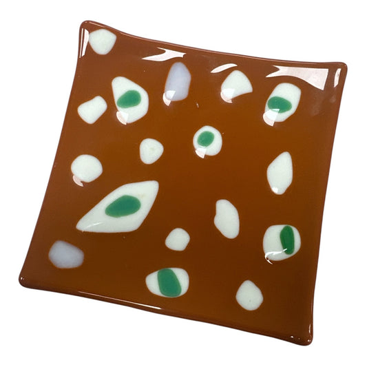 This is a square, warm brown fused-glass decorative dish scattered with an organic, abstract pattern of smooth white shapes, several of which feature vibrant green centers.