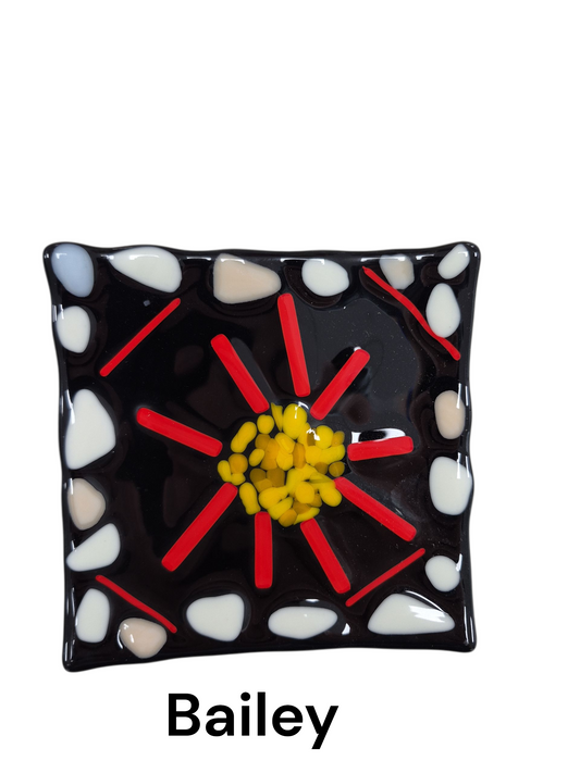 Handcrafted fused glass art tray, it features a striking, high-contrast floral motif with a textured yellow center and radiating red petals, all framed by white pebble-like accents on a sleek black background.

Medium: Fused Glass

Size: Approximately 6" x 6"

Year: 2026