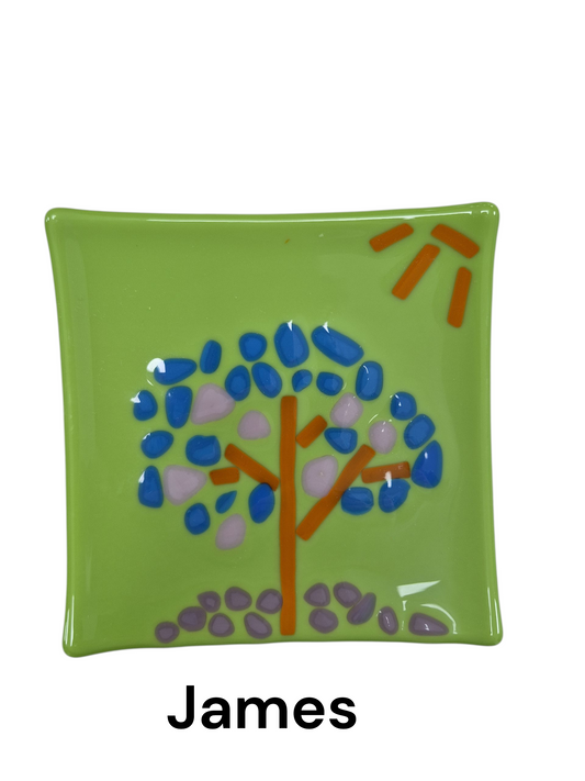 Handcrafted fused glass art tray, it showcases a whimsical, stylized tree design with a cool, wintery color palette against a vibrant, grassy-green backdrop, complete with a bright orange sun detail. To create these designs, glass artists arrange cut pieces of glass (called "frit" or "stringers") on a base piece and then heat them in a kiln until they fuse together.

Medium: Fused Glass

Size: Approximately 6" x 6"

Year: 2026