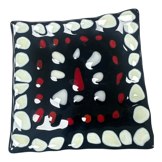 This is a square, black fused-glass decorative dish featuring a bold border of white pebble-like shapes that frame an inner square of small red accents and a central abstract arrangement of white and red glass 