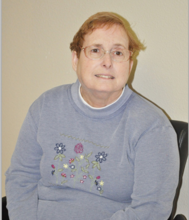 Smiling older woman with red hair and glasses, wearing a blue sweater with flowers on it.