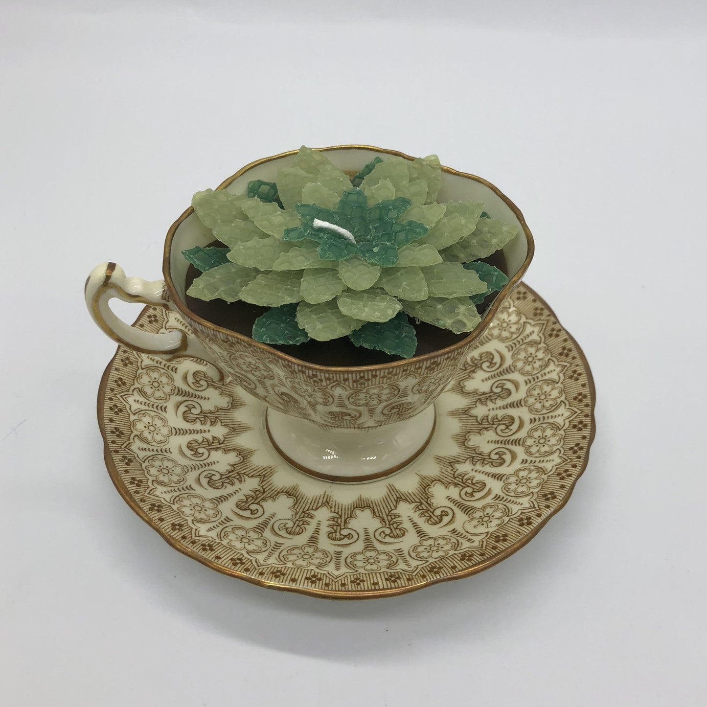 Fancy teacup with gold decorations filled with brown candle and decorated with succulent in shades of green.