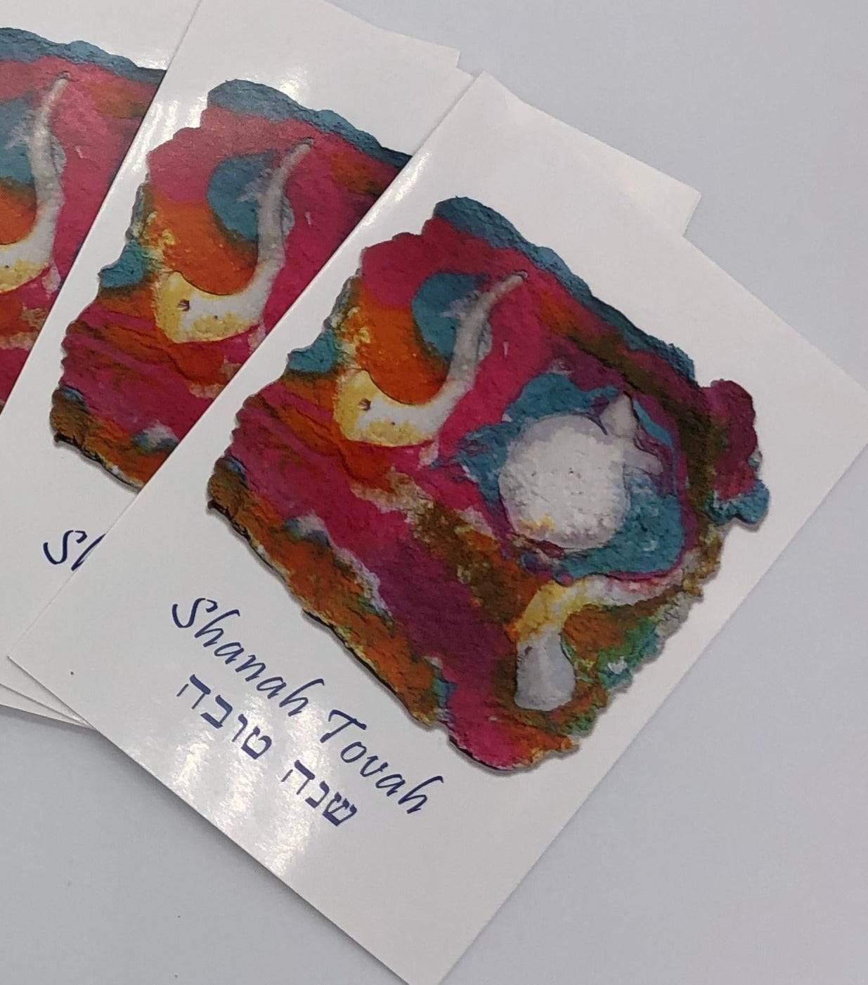 Graphic cards depicting original artwork with teal, pink and orange tones with white shofars above blue writing with both English and Hebrew Shanah Tovah