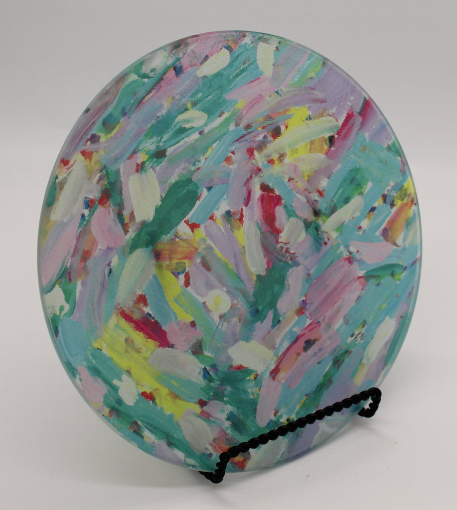 Tilted view of circular glass cutting board depicting pink, turquoise, lavender, green and yellow paint streak design