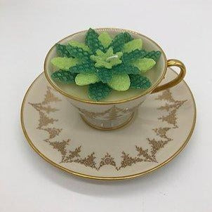 Ivory tea cup with gold edging with a light and dark green flower candle inside atop an ivory saucer with gold edging