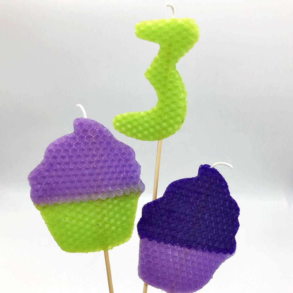 Large beeswax candles depicting a purple and green cupcake, the number 3, and a light and dark purple cupcake