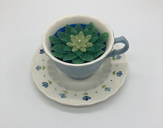 Light blue tea cup with a dark and light green flower candle inside atop a white saucer with small blue flowers
