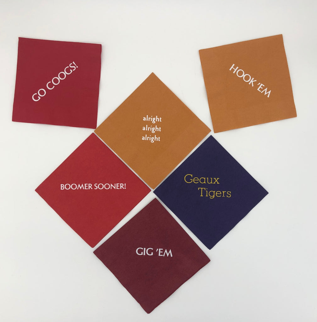 Varying colored napkins with college slogans including red napkin with Go Coogs! slogan, burnt orange napkins with Hook 'Em and alright, alright, alright slogans., purple napkin with gold Geaux Tigers slogan, red napkin with white Boomer Sooner! slogan, and maroon napkin with white Gig 'Em slogan