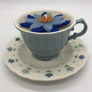 Light blue tea cup with a dark and light blue flower candle inside atop a white saucer with small blue flowers