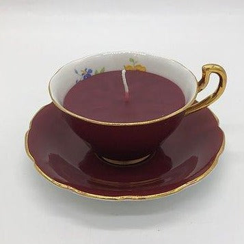 Maroon teacup with gold edging with a maroon candle inside on top of a maroon saucer with gold edging