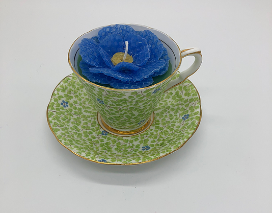 White teacup with green leaves and gold edges with a blue flower candle inside atop a white saucer with green leaves and gold edging