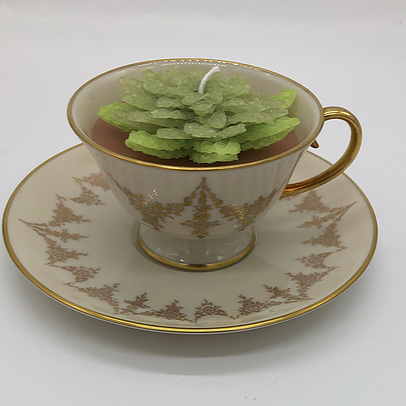 Ivory tea cup with gold edging with a light green flower candle inside atop an ivory saucer with gold edging