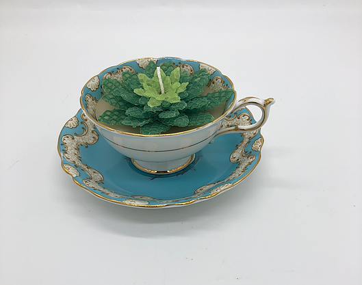 White tea cup with gold edging with a dark and light green candle inside atop a light blue saucer with gold edging