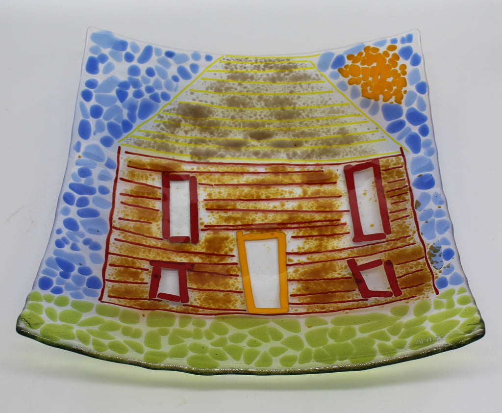 This is a square glass piece of art depicting a brown house with a blue sky and green grass.