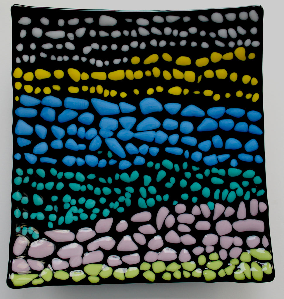 A black square glass curved plate that has small pebbles of colors. There are on average of three rows each color before transitioning to the next color. Colored pebbles are placed in the following order: white, yellow, blue, turquoise, light purple, and green.