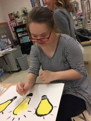 Smiling young woman wearing black and white striped shirt and red glasses painting three yellow lightbulbs