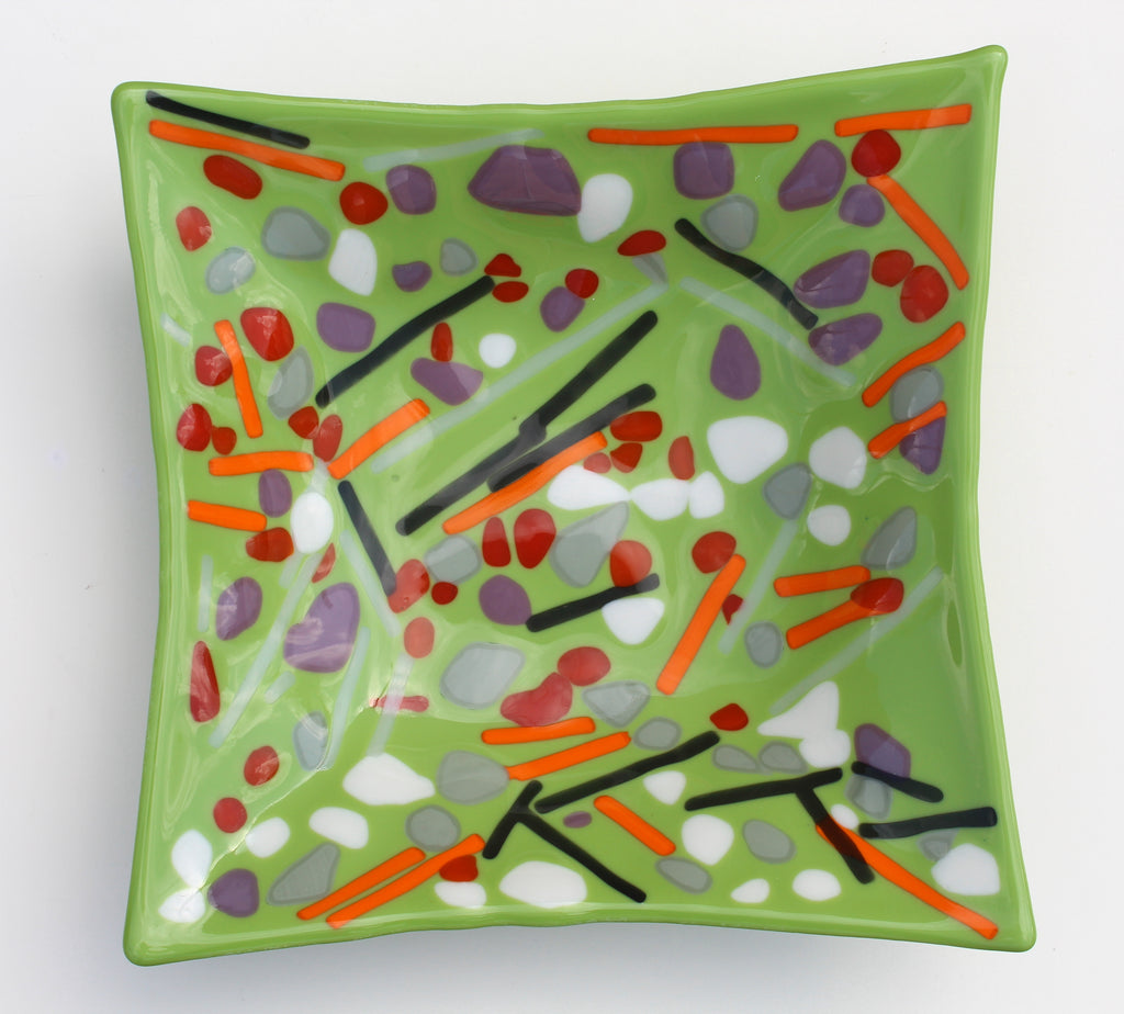 This is a fused glass piece with a green background. there are purlpe, red and white dots as well as orange and black abstract lines