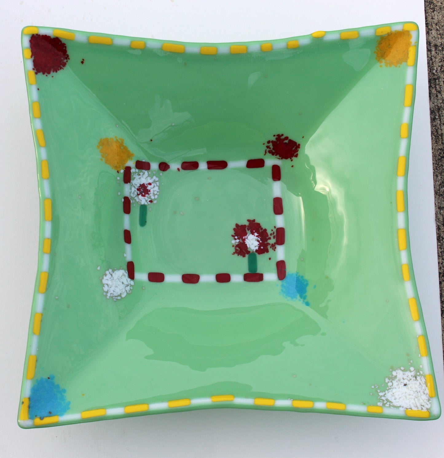 This is green fused glass with a yellow and white border, with splatters of color in each corner. COlors include yellow, burgundy, blue and white