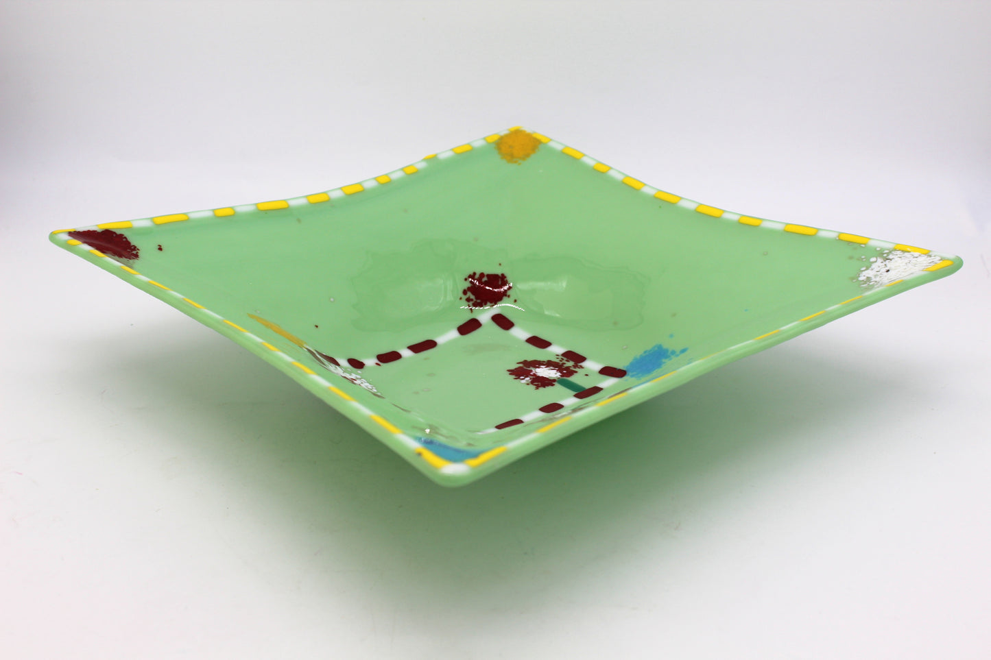 This is green fused glass with a yellow and white border, with splatters of color in each corner. COlors include yellow, burgundy, blue and white